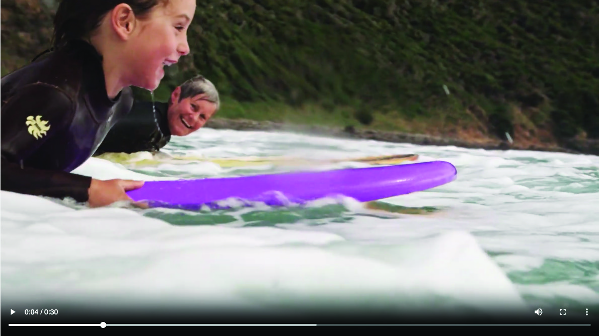 Still from the Kin video ad. Image is of a child and an older person on surf boards in a foamy body of water with a grassy cliff face behind them